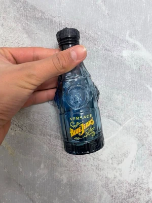 Versace Blue Jeans parfymflaska - Cool parfymflaska från Versace Blue Jeans Man. Flaskan är blå och gjord av glas med detaljerade mönster och gul text. Designen är inspirerad av retro och sticker ut i badrumshyllan. Perfekt för dig som gillar klassiska dofter och snygg design.