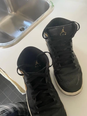 Svarta och guld Air Jordan 1 High - Säljer ett par Air Jordan 1 High sneakers i svart och guld. Skorna har klassisk hög siluett, svart läder med guldiga detaljer på swoosh, häl och logga. Tjock vit sula och svarta skosnören. Perfekta för dig som vill sticka ut med stilrena och ikoniska sneakers.