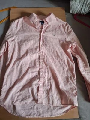 Ljusrosa skjorta från GANT - Säljer en ljusrosa skjorta från GANT med klassisk button-down krage och broderad logga på bröstet. Skjortan är långärmad och har vita knappar framtill. Materialet är bomull och passformen är normal.