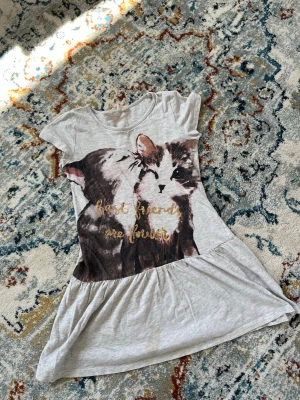 Grå klänning med kattmotiv - Söt grå kortklänning med tryck av två katter och texten 'best friends are forever' i glittriga bokstäver. Klänningen har rund halsringning och korta ärmar, samt en lätt utställd kjol. Perfekt för dig som älskar djur och vill ha en lekfull stil.
