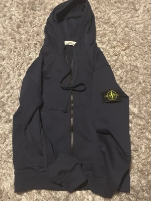 Marinblå hoodie från Stone Island - Snygg marinblå hoodie från Stone Island med dragkedja och klassisk logotyp-patch på ärmen. Huva med dragsko och mjukt material som känns skönt mot huden. Perfekt för en avslappnad streetwear-look.