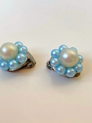 Blå pärlörhängen med retrostil - Unika örhängen med clip-on fäste, dekorerade med en stor vit pärla i mitten och omgivna av ljusblå pärlor. De har en rund form och en vintage känsla som verkligen sticker ut. Perfekta för dig som gillar att addera färg och retrovibe till din look.