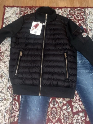 Svart pufferjacka från Moncler - Snygg svart pufferjacka från Moncler med quiltad front och stickade ärmar. Jackan har två dragkedjefickor fram, hög krage och en Moncler-logga på ärmen. Materialmix med dunfylld kropp och ribbade muddar för extra komfort.