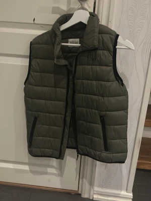 Olivgrön dunväst från Kappahl - Snygg olivgrön dunväst från Kappahl med hög krage och svarta dragkedjor på fickorna. Västen har quiltad design och är perfekt för lager-på-lager. Materialet är lätt och vadderat för extra värme. Passar till många olika stilar.