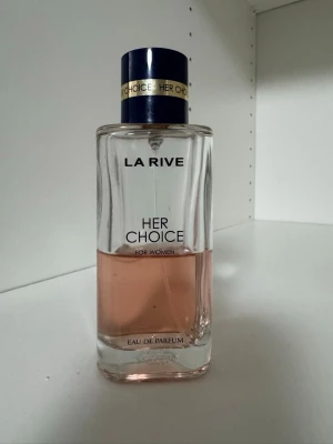 La Rive Her Choice Eau de Parfum - La Rive Her Choice Eau de Parfum för kvinnor i en elegant, fyrkantig glasflaska med ljusrosa doftvätska. Flaskan har en mörkblå kork med gulddetaljer och tydlig märkning på framsidan. Perfekt för dig som gillar fräscha och feminina dofter.