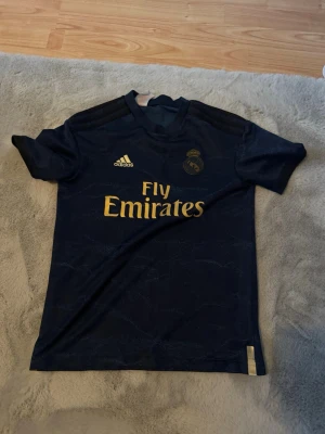 Real Madrid bortatröja Adidas blå - Mörkblå Real Madrid bortatröja från Adidas med guldiga detaljer och loggor. Tröjan har korta ärmar, rund hals och tryck med Fly Emirates på bröstet. Materialet är lätt och andas, perfekt för fotboll eller träning.