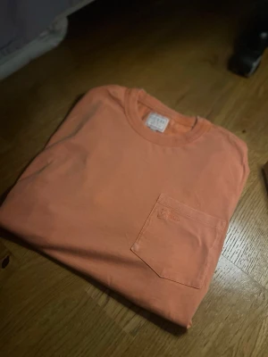 Orange t-shirt med ficka från GUESS - Snygg orange t-shirt från GUESS med rund halsringning och en bröstficka där loggan är broderad ton-i-ton. Tillverkad i mjuk bomull och har en klassisk passform som funkar till det mesta. Perfekt för dig som gillar enkel men stilren design.