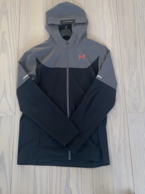 Svart och grå vindjacka Under Armour - Snygg vindjacka från Under Armour i svart och grått med hel dragkedja och huva. Jackan har en röd logga på bröstet och reflekterande detaljer på ärmarna. Perfekt för blåsiga dagar och sportiga outfits.
