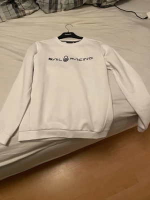 Vit sweatshirt från Sail Racing - Vit sweatshirt från Sail Racing med svart logotyptryck på bröstet. Tröjan har rund halsringning med svartvit randig detalj på insidan och är tillverkad i mjukt material. Perfekt för en stilren och sportig look.
