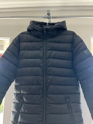 Svart dunjacka med huva - Svart dunjacka med huva och quiltad design. Jackan har två fickor med dragkedja och en patch på ena ärmen. Perfekt för kalla vinterdagar och har en stilren look som passar till det mesta.