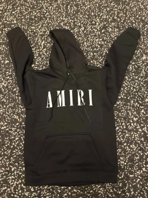 Svart hoodie från Amiri med tryck - Svart hoodie från Amiri med stort vitt logotryck på bröstet. Klassisk modell med huva och dragsnören, samt en stor magficka framtill. Materialet ser ut att vara mjukt och bekvämt, perfekt för en avslappnad och cool look. Passar också storlek M eftersom den är liten i storlek