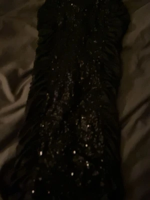 Svart glittrig klänning från Kivenst - En svart klänning från Kivenst High Fashion med glittriga detaljer och rynkad design längs sidorna. Klänningen har en elegant och festlig look med en smal passform som framhäver siluetten. Perfekt för dig som vill sticka ut med lite extra glitter.