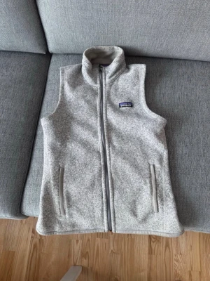 Ljusgrå fleeceväst från Patagonia - Säljer en ljusgrå fleeceväst från Patagonia med hög krage och dragkedja framtill. Västen har två fickor med dragkedja och en liten logga på bröstet. Perfekt att slänga på sig över en hoodie eller tröja för extra värme. OBS, ändast använd 3 gånger!