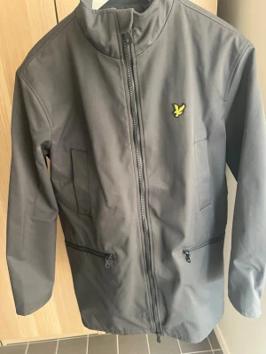 Mörkgrå Parka från Lyle & Scott - Snygg mörkgrå parka från Lyle & Scott. Jackan har hög krage, två sidofickor och två dragkedjefickor nedtill. Materialet är slätt och tåligt, perfekt för blåsiga dagar. Använd ett fåtal gånger.