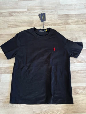 Svart t-shirt från Ralph Lauren - Klassisk svart t-shirt från Ralph Lauren med den ikoniska röda broderade polospelaren på bröstet. T-shirten har rund hals och korta ärmar. Tillverkad i mjuk bomull för en skön och avslappnad känsla.