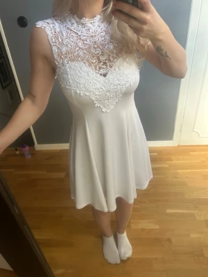 Vit spetsklänning med knyt i nacken - Säljer en vit kortklänning med vacker spets upptill och hög hals. Klänningen har ett elegant knyt i nacken och är ärmlös med en lätt utsvängd kjol. Perfekt för sommarens alla tillfällen och studentfirande.
