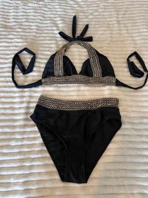 Svart bikini med mönstrad kant - Snygg svart bikini med beige och svart mönstrad kant på både överdel och underdel. Överdelen har halterneck och knyt i nacken samt ryggen. Materialet känns mjukt och stretchigt, perfekt för lata dagar på stranden.