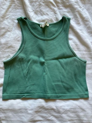 Grön ribbad croppad linnetopp - Snygg grön croppad linnetopp i ribbad bomull. Toppen är ärmlös med breda axelband och har en enkel, sportig look. Perfekt att styla med höga jeans eller shorts för en chill vibe.