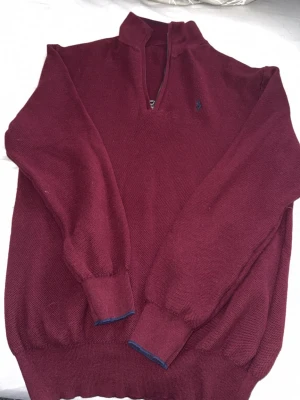Vinröd stickad tröja från Polo Ralph Lauren - Snygg vinröd stickad tröja från Polo Ralph Lauren med halv dragkedja och hög krage. Tröjan har långa ärmar, ribbade muddar och den klassiska broderade loggan på bröstet. Perfekt för dig som gillar stilrena och tidlösa plagg.