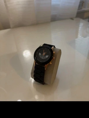 Klocka - Snygg svart klocka från Adidas med svart länkarmband och guldfärgade detaljer på boetten. Urtavlan har Adidas-loggan i silver och orange visare. Perfekt accessoar för dig som gillar sportig och stilren design.