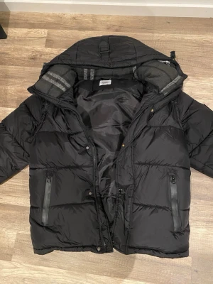 Svart pufferjacka från Burberry (Kan göras till Väst) - Snygg svart pufferjacka från Burberry med huva och dragkedja framtill. Jackan har två framfickor med dragkedja och en patch med texten 'BURBERRY LONDON ENGLAND' på ärmen. Insidan har ett klassiskt rutmönster i grått och svart. Perfekt för kalla vinterdagar.