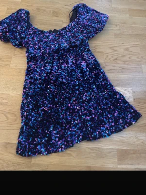 Glittrig kortklänning med puffärm - Säljer en glittrig kortklänning med puffärmar i blått, lila och rosa. Klänningen har en rundad urringning och är täckt av paljetter som ger ett riktigt partyvibe. Perfekt för dig som vill sticka ut och glänsa på dansgolvet!