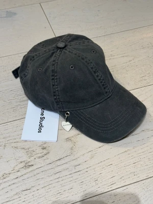 Svart keps från Acne Studios - Snygg svart keps från Acne Studios i tvättad bomull med böjd skärm och diskret logga broderad på sidan av skärmen. Kepsen har metallhål för ventilation och ett litet hjärtformat metallhänge med Acne Studios ingraverat. Perfekt accessoar för en trendig look.