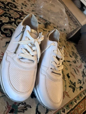 Vita sneakers med snörning och dragkedja - Snygga vita sneakers med perforerat mönster framtill, snörning och dragkedja på sidan. Skorna har en sportig och modern look med detaljer i vitt och silver. Perfekta för dig som gillar stilrena och fräscha sneakers.