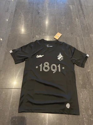 Svart AIK tröja från Nike - Snygg svart AIK t-shirt från Nike med tryck '1891' på bröstet och klubbens emblem. Tröjan har rund hals, korta ärmar och är tillverkad i ett lätt och funktionellt material, perfekt för träning eller match. Har i alla storlekar 