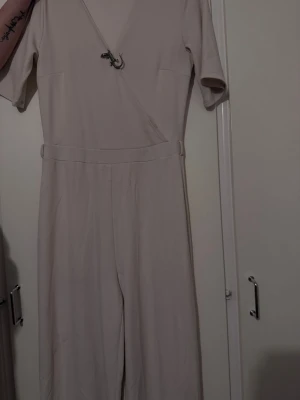 Beige kortärmad jumpsuit med omlott - Snygg beige jumpsuit med korta ärmar och omlott-ringning. Modellen har markerad midja med bälte och vida ben. Perfekt för dig som gillar en stilren och enkel look. Materialet känns mjukt och följsamt mot kroppen.