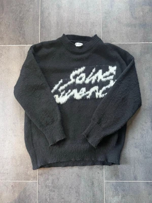 Svart Saint Laurent stickad tröja -  svart stickad tröja från Saint Laurent med vit fuzzy logotyp på bröstet. Tröjan har rund halsringning, ribbade muddar och är tillverkad i ett mjukt stickat material som känns lyxigt och trendigt.