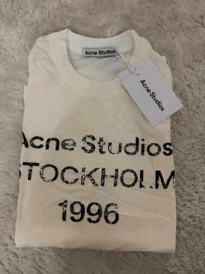 Vit Acne Studios Tröja Logga - Säljer en vit t-shirt från Acne Studios med tryck 'Stockholm 1996' framtill, långärmad. Perfekt basic-plagg med cool print för en avslappnad stil. Helt ny med prislapp, kvitto finns. Nypris 3500kr