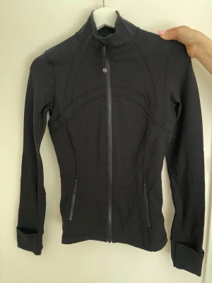 Svart träningsjacka från Lululemon - Säljer en svart träningsjacka från Lululemon med figurnära passform och hög krage. Jackan har långa ärmar med tumhål, två fickor med dragkedja och snygga sömdetaljer som ger en sportig look. Perfekt för gymmet eller löprundan. I deras storlekssystem motsvarar det ungefär:  * XS / Small * EU ca 34–36