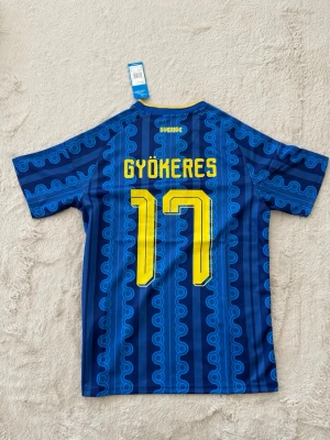 Sverige fotbollströja Gyökeres #17 - Blå fotbollströja från Adidas med Sveriges landslagsemblem, Gyökeres och nummer 17 i gult. Tröjan har mönstrade blå ränder, gula detaljer och klassiska Adidas-ränder på axlarna. Tillverkad i lätt funktionsmaterial med V-ringning.