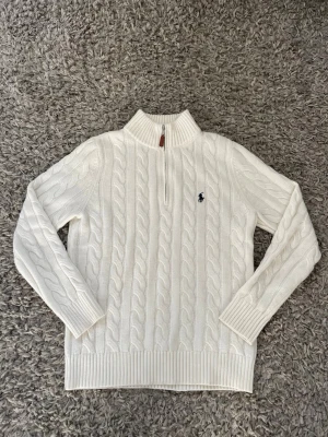 Vit kabelstickad tröja Polo Ralph Lauren - Kabelstickad vit tröja från Polo Ralph Lauren med halv dragkedja och ribbad krage. Tröjan har det klassiska blå logobroderiet på bröstet och är tillverkad i mjukt material. Perfekt för en clean och stilren look.