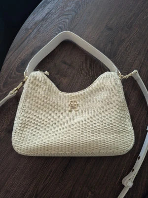 Beige flätad handväska Tommy hilfiger  - Snygg beige handväska från Tommy hilfiger med flätad design och vit axelrem i läder. Väskan har ett guldfärgat logomärke på framsidan och stilrena detaljer. Den har några små märken, se bild. Men inget som direkt syns. Samt några små fläckar på insidan. Annars i fint skick. Det långa bandet är avtagbar