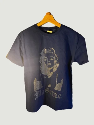 Blå t-shirt med tryck från Masteme - Cool blå t-shirt från Masteme med ett stort glittrigt tryck framtill föreställande ett ansikte och texten 'masteme'. Klassisk passform och rund hals. Perfekt för dig som vill sticka ut med en unik design.