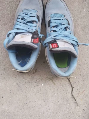 Jordan 4 Retro University Blue sneakers - Säljer ett par Jordan 4 Retro University Blue sneakers med ljusblå mocka, svarta och vita detaljer samt meshpaneler. Skorna har klassiska '23'-patches på tungan och signaturdetaljer. Perfekt för dig som vill sticka ut med ikoniska sneakers.