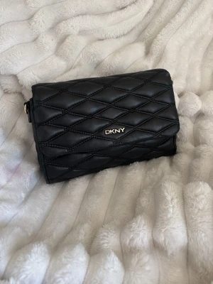 Axelväska från DKNY - Snygg svart axelväska från DKNY i quiltat mönster. Väskan har guldfärgade detaljer och en axelrem i kedja med inslag av svart läder. Perfekt storlek för det viktigaste och stängs med lock. Klassisk och trendig design som lyfter vilken outfit som helst. Lite slitet men inget man tänker på (därav priset) 