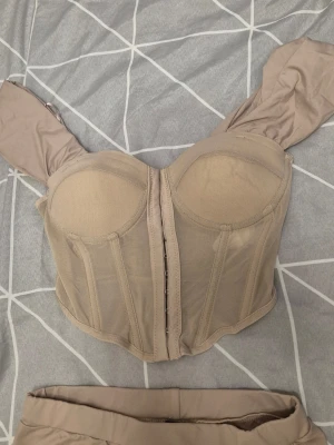 Beige mesh bustier och kjol set - Snyggt set med beige bustier i mesh med vadderade kupor, axelband och markerade sömmar framtill. Matchande lång kjol med omlott-design. Perfekt för dig som gillar trendiga och stilrena underkläder.