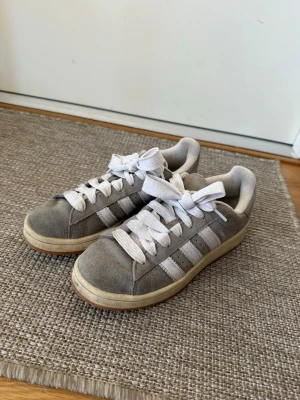 Grå Adidas sneakers med vita ränder - Snygga grå sneakers från Adidas med klassiska tre vita ränder på sidorna och vita skosnören. Ovandelen är i mockaliknande material. Perfekta för en avslappnad streetstyle-look.