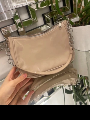 Beige axelväska med kedjedetaljer - Snygg beige axelväska i glansigt material med silverfärgad kedja som axelrem och dragkedja upptill. Väskan har en mjukt rundad form och minimalistisk design, perfekt för dig som gillar stilrena accessoarer. Ingen synlig logga eller märke.
