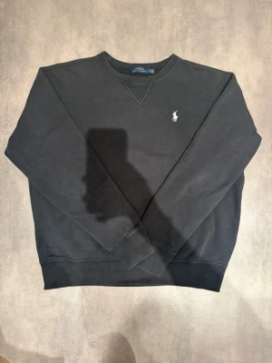 Svart sweatshirt från Polo Ralph Lauren - Klassisk svart sweatshirt från Polo Ralph Lauren med rund halsringning och broderad vit logga på bröstet. Tröjan har ribbade muddar vid ärmslut och nederkant, perfekt för en stilren och avslappnad look.