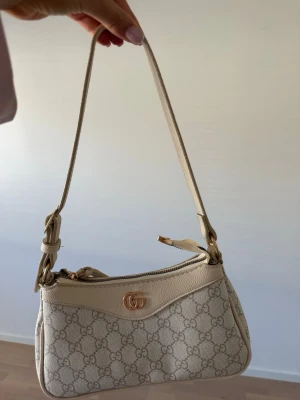 Väska - Snygg beige handväska med guldfärgad logga framtill. Väskan har axelrem, dragkedja och detaljer i läder. Perfekt för dig som gillar lyxiga accessoarer och vill sticka ut med en ikonisk design.