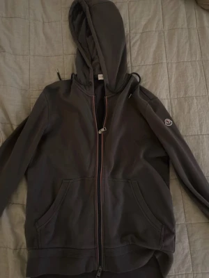 Brun hoodie från Moncler med dragkedja - Brun hoodie från Moncler med hel dragkedja framtill, huva med dragsko och två stora fickor. Klassisk Moncler-logga på ärmen och ribbade muddar. Perfekt för en avslappnad och trendig stil.