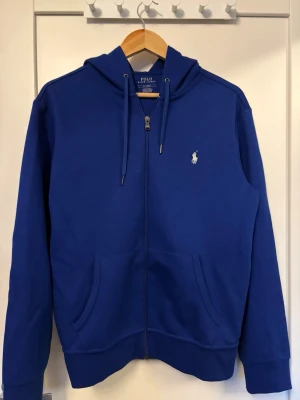 Blå hoodie från Polo Ralph Lauren - Snygg blå hoodie från Polo Ralph Lauren med dragkedja framtill, två fickor och klassisk huva med dragsko. Ikoniska loggan broderad på bröstet. Tillverkad i mjuk bomullsblandning som känns skön mot huden. Perfekt för en avslappnad och stilren look.🙌🤩👔
