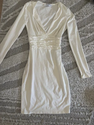 Vit studentklänning - Säljer en  vit kortklänning från By Swan med omlott-ringning och långa ärmar. Klänningen har markerad midja med rynkade detaljer. Perfekt som studentklänning eller sommar/ midsommar klänning.