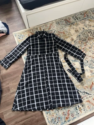 Svartvit rutig skjortklänning - Svart skjortklänning med vitt rutmönster och knappar längs hela framsidan. Klänningen har lång ärm, klassisk krage och avtagbart bälte i midjan. Perfekt för dig som gillar stilrena och trendiga plagg.