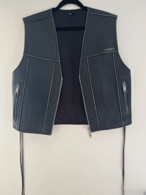 Svart skinnväst från EYTYS - Snygg svart skinnväst med silverfärgade dragkedjor och detaljer. Västen har två fickor med dragkedja och dekorativa sömmar. Perfekt för att lyfta din outfit med en edgy touch.