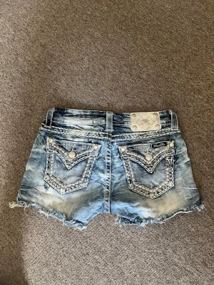 Blå jeansshorts från Miss Me - Säljer ett par blå jeansshorts från Miss Me med slitna kanter och snygga detaljerade bakfickor med knappar och broderi. Klassisk femficksmodell med låg midja och cool tvättad look. Perfekta för sommaren!